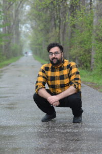 IMAGE DESCRIPTION: Outdoors Mohammad Talha Akbar crouches on a tree-lined path while looking at the camera.