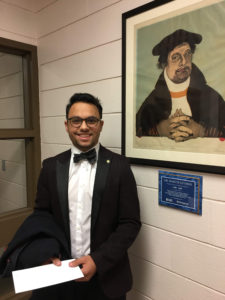 Kiegan Lloyd (he/him) Luther Students Director wearing a black suit with a white shirt, black bow tie, and a smile, stands near a painting pf Dr. Martin Luther.