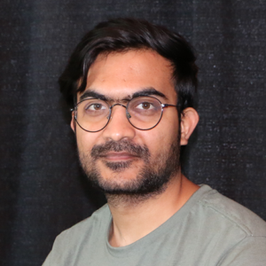 Udaykumar Posia (he/him) Junior Developer poses for his staff photo. Behind him is a black curtain.