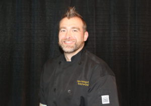 Ryan Katchuk (he/him) URSU Kitchen Manager sits in front of a black curtain for a staff photo. He is wearing a black chef's jacket with his name and Executive Chef on it.