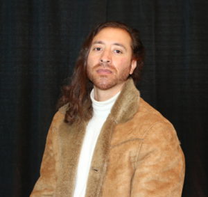 Edgar Martinez (he/him) URSU Graphic Designer poses for his staff photo. His luxurious hair falls over his right shoulder, hi is wearing a stylish tan suede jacket and white turtleneck.